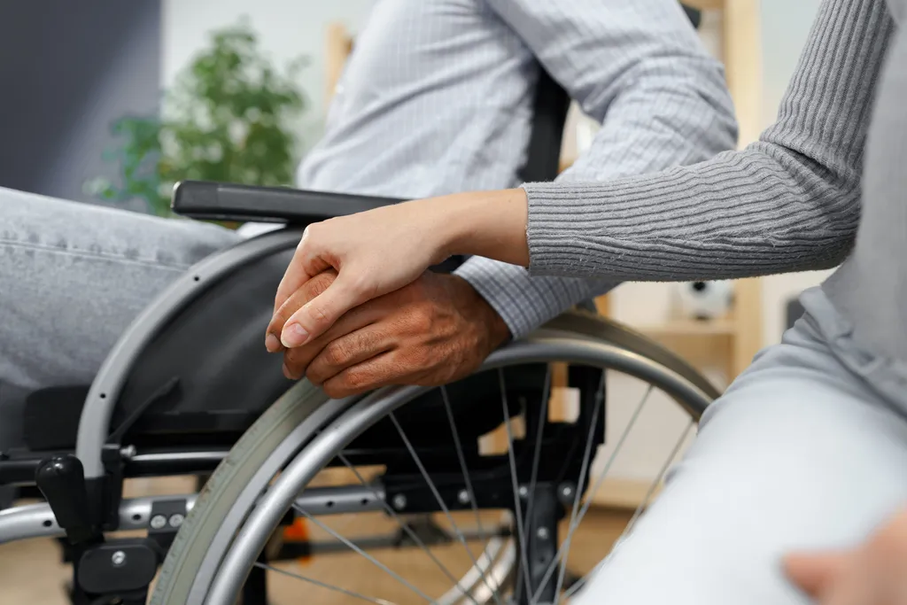 Pflegekraft legt ihre Hand auf die Hand eines Mannes im Rollstuhl